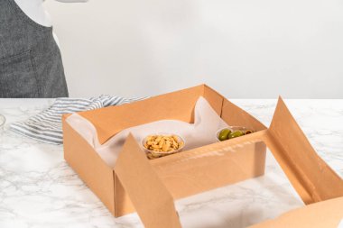 Kurulum sürecinde, bir kadın bu şarküteri kutusunu ustalıkla düzenliyor, dilimlenmiş et, peynir, kraker ve üzüm sergiliyor, hepsi düzgünce düzenlenmiş ve kahverengi bir hediye kutusunda paketlenmiş..