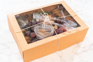 Dilimlenmiş et, peynir, kraker ve üzümlerden oluşan şarküteri kutusu, hepsi kahverengi bir hediye kutusunda güzelce paketlenmiş..