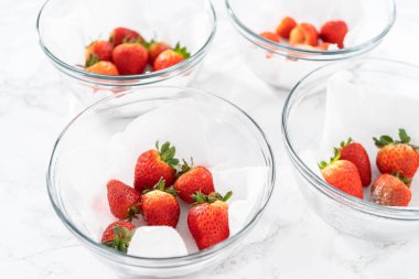 Parlak kırmızı çilekler, küf izleriyle karışmış, beyaz peçetedeki kağıt havluyla kaplı cam bir kasede dinleniyor, depolama tekniklerini gösteriyor..