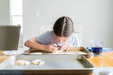 Küçük bir kızın özenle yazdığı iç açıcı bir sahne. Şekerli kurabiyelerin üzerine besin boyası sürmüş. Kurabiyeler güzel bir şekilde beyaz kraliyet kreması ile dolmuş..