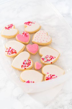 Kalp şeklinde şeker kurabiyelerini büyük plastik bir kapta pembe ve beyaz kraliyet kreması ile depolamak.