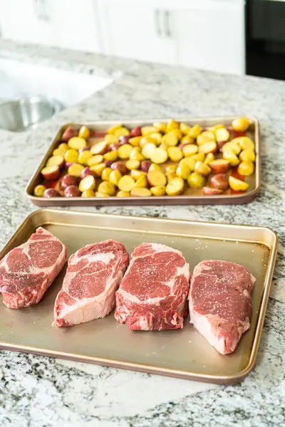 Situated in a modern white kitchen, a seasoned rib eye steak, boasting its beautiful marbling, sits ready on a baking sheet. It is prepared for the outdoor gas grill, promising a perfect sear on the