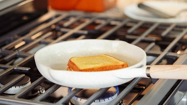 Within the sleek setting of a modern white kitchen, banana slices and brioche bread sizzle on a non-stick pan over a gas stove, setting the stage for an enticing grilled peanut butter banana sandwich.