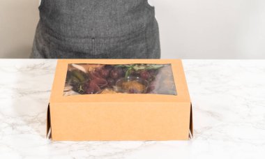 Dilimlenmiş et, peynir, kraker ve üzümlerden oluşan şarküteri kutusu, hepsi kahverengi bir hediye kutusunda güzelce paketlenmiş..