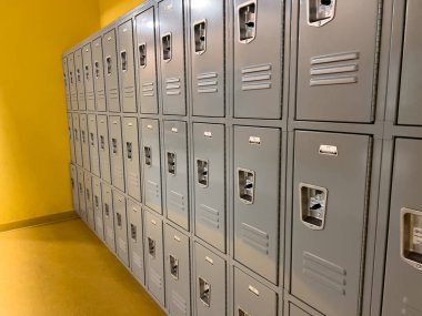 Bir okul koridorunda, öğrenciler için gri dolaplar dizili, havalandırmalar ve kilitler pürüzsüz metal kapıları noktalıyor..