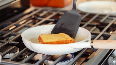 Modern beyaz bir mutfak, muz dilimleri ve brioche ekmek cızırtısı içinde gaz sobasının üzerinde, baştan çıkarıcı bir ızgara fıstık ezmeli muzlu sandviç için zemin hazırlıyor..