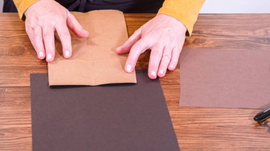 Adım adım. Öğretmen, kahverengi bir çantadan kağıt bir kukla yaparak, çalışma alanı olarak ahşap bir yüzey kullanarak çevrimiçi sınıfa rehberlik ediyor..