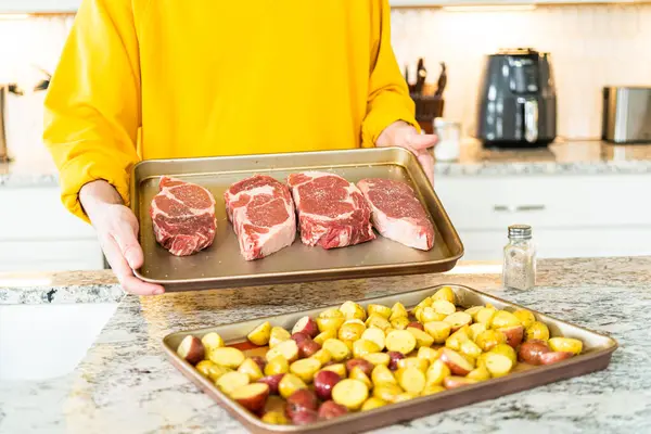 Situated in a modern white kitchen, a seasoned rib eye steak, boasting its beautiful marbling, sits ready on a baking sheet. It is prepared for the outdoor gas grill, promising a perfect sear on the