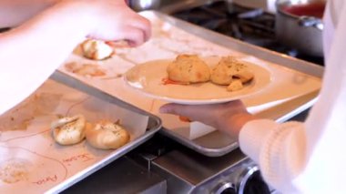 Altın kahverengisi mini calzones, otlarla süslenmiş, beyaz bir tabakta oturuyor, fırından yeni çıkmış. Arka planda daha fazla calzone ile dolu bir pişirme çarşafı var, hepsi de şenlikli bir yemeğin parçası.