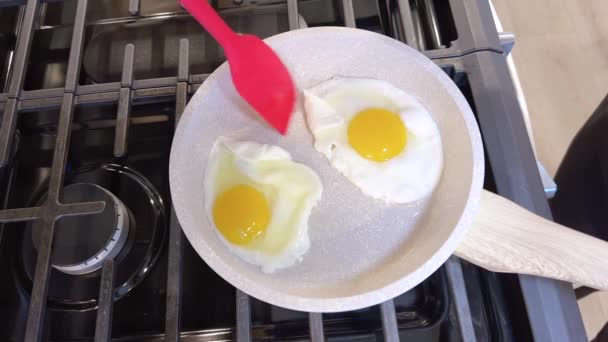 两个煎得恰到好处的阳光普照的鸡蛋放在一个不粘的煎锅里 上面放着一个红色的硅胶铲 准备上菜 全部放在炉顶上 — 图库视频影像