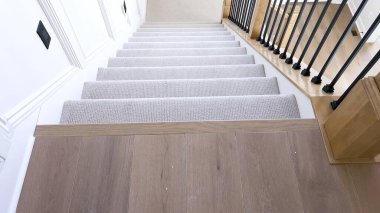 Görüntü, beyaz duvarlar ve ahşap balyalarla tamamlanmış pelüş bej halıyla kaplı iyi tasarlanmış modern bir merdivenin detaylarını yakalıyor..