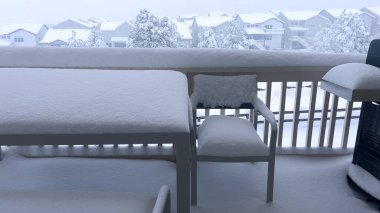 Kalın kar tabakaları dış mobilyalarla kaplı bir balkon, kışın örtülü banliyö manzarasını huzurlu bir şekilde sunuyor..
