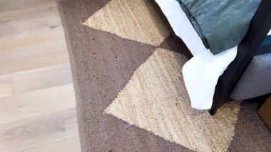 Modern bir yatak odasının köşesinde şık bir geometrik halı ve parlak, çağdaş mobilyalar bulunur. Pürüzsüz, bronz bir şilte yatakları, temiz çizgileri ve nötr renk paletini tamamlar..