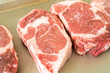 Modern beyaz bir mutfakta, baharatlı bir pirzola bifteği, güzel mermerleriyle övünerek, bir pişirme çarşafının üzerinde oturur. Açık hava gaz ızgarası için hazırlanıyor, mükemmel bir kavurucu söz veriyor.