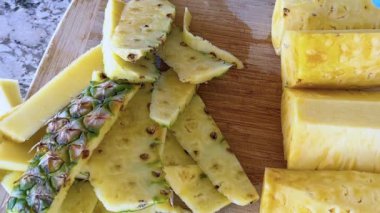 Hijyen için mavi tek kullanımlık eldivenler giyen biri, temiz mutfak tezgahına kurulmuş, ahşap bir kesme tahtasındaki sulu organik ananası ustalıkla dilimliyor..