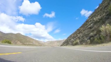 Araç, parlak güneşin altında Cuyama Otoyolu boyunca ilerliyor. Çevresindeki manzara, ışık saçan güneş ışığıyla aydınlanır ve araba seyahat ederken güzel ve davetkar bir sahne yaratır.