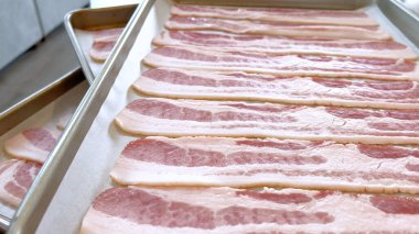 Pişirme tepsisine yerleştirilmiş çiğ pastırma şeritleri, bu popüler kahvaltı malzemesinin çıtır çıtır ve altın rengine dönüşmeden önce taze ve pişmemiş görüntüsünü yakalıyor..