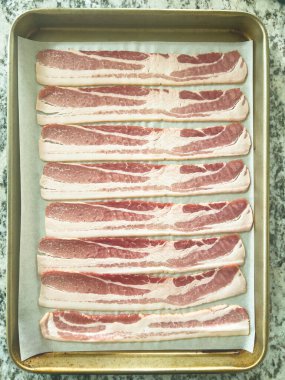 Pişirme tepsisine yerleştirilmiş çiğ pastırma şeritleri, bu popüler kahvaltı malzemesinin çıtır çıtır ve altın rengine dönüşmeden önce taze ve pişmemiş görüntüsünü yakalıyor..