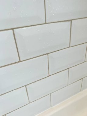 Beyaz fayanslı bir duş duvarının yakın çekimi, seramiklerin düzgün hizalanmasını ve su damlacıklarının varlığını gösteriyor. Bu resim bir duş alanının temiz ve modern tasarımını vurgular,