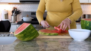 Sarı kazaklı bir kadın modern bir mutfakta taze bir karpuzu kesme tahtasıyla dilimliyor. Mutfakta beyaz dolap, altıgen döşeme ve çeşitli mutfak aletleri var.