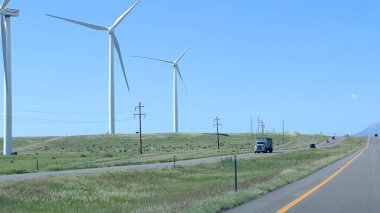 Rüzgâr türbinleri, açık mavi gökyüzünün altındaki yeşil tarlaları ve uzak kar kaplı dağları olan Colorado 'da bir otoyol boyunca dimdik dururlar. Arabalar ve kamyonlar yolda görülebilir.