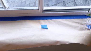 Titizlikle maskelenmiş kenarlar ve örtülü yüzeyler, konut penceresinin pervazını boyamak için atılan ilk adımları ortaya koyuyor. Mavi bandın kumlu bej kumaşa karşı olan kontrastı,