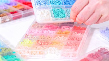 Kadın elleri şeffaf organizatörlerin renklerine göre sıralanmış boncuk koleksiyonunun üzerinde zarif bir şekilde duruyor. Boncuk dizisi, derin morlardan parlak portakallara kadar, titizlikle geniş bir yelpazeyi kapsıyor.