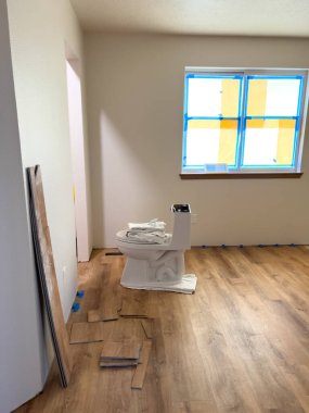 Görüntü, yenilenmekte olan bir odada kısmen tamamlanmış ahşap zemin kurulumunu gösteriyor. Bir tuvalet, inşaat malzemeleri ve aletlerle çevrili olarak geçici olarak odada durur. İlerleme...