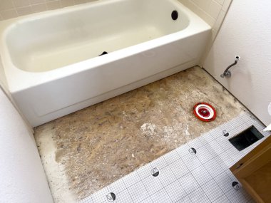 Görüntü, bir yenileme projesinin ortasında bir banyo sergiliyor. Açığa çıkmış bir zemin ve kısmen döşenmiş yeni döşeme hazırlıkları var. Küvet bozulmamış.