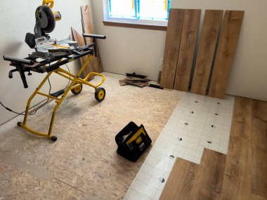 Görüntü, tadilattan geçen bir odada kullanılmaya hazır sarı bir tezgaha yerleştirilmiş bir miter testeresini gösteriyor. Kısmen döşenmiş ahşap zemin ve inşaat malzemeleri devam eden döşeme çalışmalarını gösteriyor. Şey...