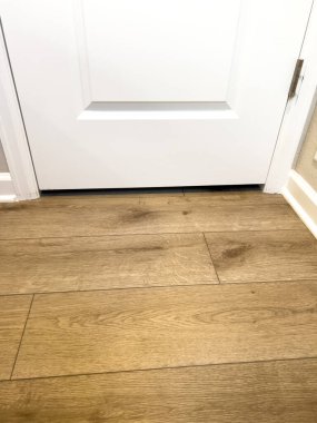 This image features a close-up view of a section of newly installed wood flooring, highlighting the area at the base of a white door. The wood flooring has a light, natural finish with visible wood clipart