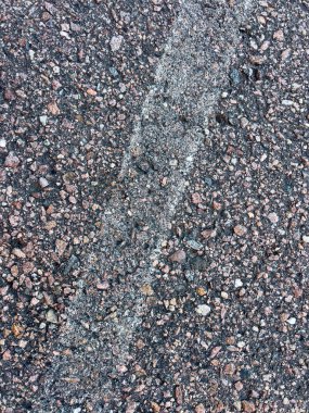 Soluk beyaz çizgi işaretli asfalt bir yol yüzeyine yakın çekim. Asfaltın kaba dokusu, içine yerleştirilmiş küçük taşlar ve çakıl taşlarının karışımı ile vurgulanır ve aşınma izleri gösterir.