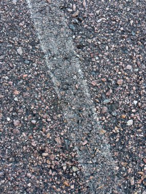 Soluk beyaz çizgi işaretli asfalt bir yol yüzeyine yakın çekim. Asfaltın kaba dokusu, içine yerleştirilmiş küçük taşlar ve çakıl taşlarının karışımı ile vurgulanır ve aşınma izleri gösterir.