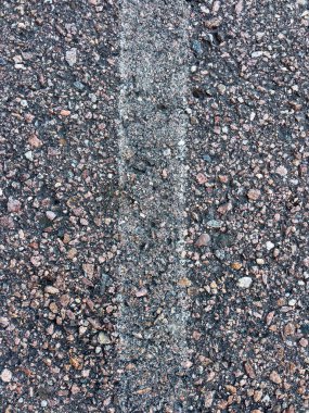 Soluk beyaz çizgi işaretli asfalt bir yol yüzeyine yakın çekim. Asfaltın kaba dokusu, içine yerleştirilmiş küçük taşlar ve çakıl taşlarının karışımı ile vurgulanır ve aşınma izleri gösterir.