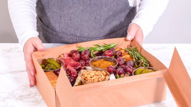 Elleri titizlikle bir kaseye taze kırmızı üzüm ekliyor, çeşitli peynirler, zeytinler ve pişirilmiş etlerle dolu güzel bir şarküteri kutusunu tamamlıyor.