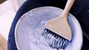 Zengin, kremsi saç boyası süslü bir kasenin üzerindeki ahşap fırçanın üzerinde duruyor. Saç boyama araçları koyu, dokulu bir havluya uzanır. İçinden canlı pembe bir işaret geçer.