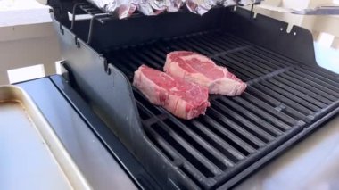 Bu resim barbekü ızgarasında pişen üç kalın bifteği, yukarıdaki koçanda folyoya sarılı mısırla birlikte, tipik bir açık hava yemek sahnesini görüntülüyor.