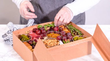 Elleri titizlikle bir kaseye taze kırmızı üzüm ekliyor, çeşitli peynirler, zeytinler ve pişirilmiş etlerle dolu güzel bir şarküteri kutusunu tamamlıyor.