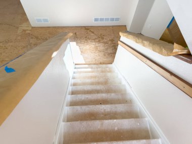 Üst kattaki bir koridorda çıplak zemin ve ahşap korkuluklardan oluşan bir merdiven manzarası. Güneş ışığı, ev yenileme projesinde kaydedilen ilerlemeyi göstererek alanı aydınlatır.