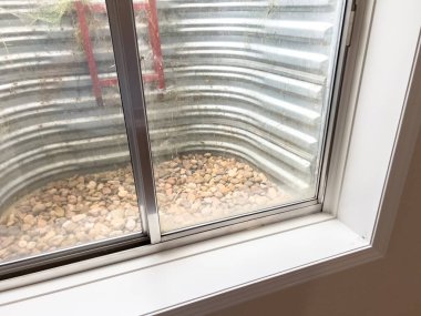 Bodrum penceresinin yakın plan görüntüsü beyaz iç pencere çerçevesini gösteriyor. Metal bir pencerenin dış görünüşü ise küçük taşlarla kaplı. Temiz pencere çerçevesi ve iyi korunmuş çerçeve