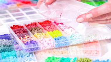 Kadın elleri şeffaf organizatörlerin renklerine göre sıralanmış boncuk koleksiyonunun üzerinde zarif bir şekilde duruyor. Boncuk dizisi, derin morlardan parlak portakallara kadar, titizlikle geniş bir yelpazeyi kapsıyor.