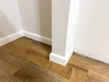 Beyaz vinil süpürgelikler ve süsleme parçaları, ahşap döşemeli bir odaya yerleştirilmek üzere bir duvara karşı düzgünce dizilmiş. Yenileme projesi devam ediyor, odayı tamamlamaya hazır.