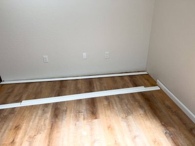 Taze ahşap döşemeli, iki pencereli ve duvarlar boyunca yerleştirilmeye hazır beyaz vinil süpürgelikli bir oda. Alan parlak ve yenileme sürecinde ilerleme gösteriyor.