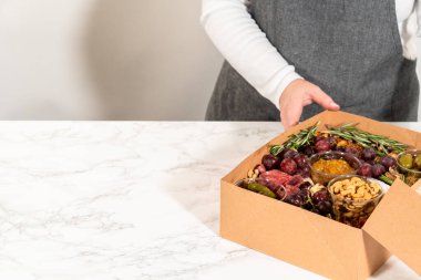 Dilimlenmiş et, peynir, kraker ve üzümlerden oluşan şarküteri kutusu, hepsi kahverengi bir hediye kutusunda güzelce paketlenmiş..