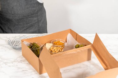 Kurulum sürecinde, bir kadın bu şarküteri kutusunu ustalıkla düzenliyor, dilimlenmiş et, peynir, kraker ve üzüm sergiliyor, hepsi düzgünce düzenlenmiş ve kahverengi bir hediye kutusunda paketlenmiş..