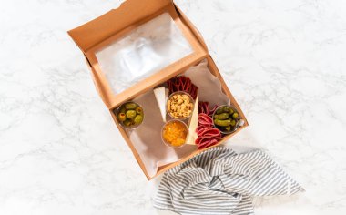 Düz yatıyordu. Kurulum sürecinde, bir kadın bu şarküteri kutusunu ustalıkla düzenliyor, dilimlenmiş et, peynir, kraker ve üzüm sergiliyor, hepsi düzgünce düzenlenmiş ve kahverengi bir hediye kutusunda paketlenmiş..