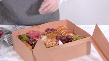 Elleri titizlikle bir kaseye taze kırmızı üzüm ekliyor, çeşitli peynirler, zeytinler ve pişirilmiş etlerle dolu güzel bir şarküteri kutusunu tamamlıyor.