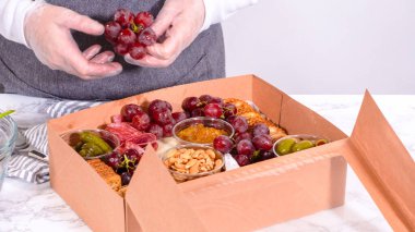 Elleri titizlikle bir kaseye taze kırmızı üzüm ekliyor, çeşitli peynirler, zeytinler ve pişirilmiş etlerle dolu güzel bir şarküteri kutusunu tamamlıyor.