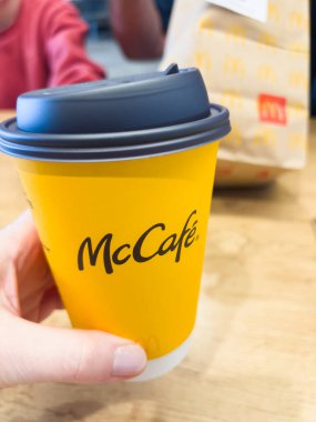St. George, Utah, 10 Kasım 2024. McDonald 'daki ahşap bir masaya yerleştirilmiş koyu gri kapaklı sarı bir McCafe kahve fincanını tutan bir el. Arka planda bir McDonald kahvaltı çantası var.
