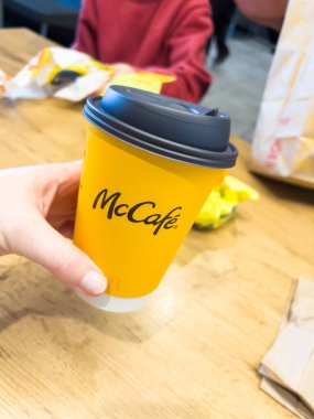 St. George, Utah, 10 Kasım 2024. McDonald 'daki ahşap bir masaya yerleştirilmiş koyu gri kapaklı sarı bir McCafe kahve fincanını tutan bir el. Arka planda bir McDonald kahvaltı çantası var.